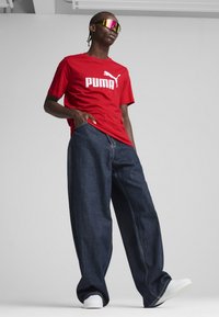 Rotes T-Shirt mit kurzen Ärmeln und weißem "PUMA"-Logo, kombiniert mit lockeren, dunklen Jeans und weißen Turnschuhen. Das Modell trägt reflektierende Sonnenbrillen.