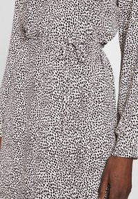 Šaty s dlouhým rukávem z lehké látky s béžovým podkladem a černým puntíkovaným vzorem, s nastavitelným páskem v pase a manžetami na rukávech.