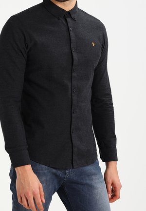 Homme portant une chemise noire ajustée à manches longues boutonnée avec un petit logo jaune sur la poitrine gauche et un jean bleu.