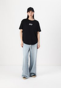 Svart bomulls-t-skjorte med "GAP"-logo, kombinert med lyseblå flared jeans. Modellen har på seg en svart caps og beige sneakers.