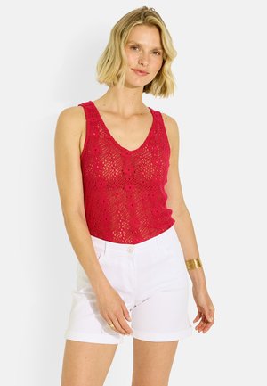 Mujer con cabello rubio corto, vestida con un top rojo sin mangas de crochet y unos pantalones cortos blancos de cintura alta, de pie frente a un fondo blanco.