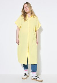 Vestido de manga corta y botón en suave algodón amarillo con un corte holgado. Presenta un corte recto y una textura sutil, combinado con jeans azules y zapatillas.