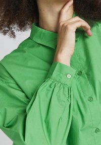 Green cotton shirt featuring a classic collar, button-up front, and gathered detailing at the sleeves. Smooth texture with matching buttons.