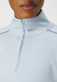 Svijetloplava majica s polu-zatvaračem napravljena od glatkog, rastezljivog materijala. Ima srebrni Nike logo na ovratniku i kontrastne šavove.