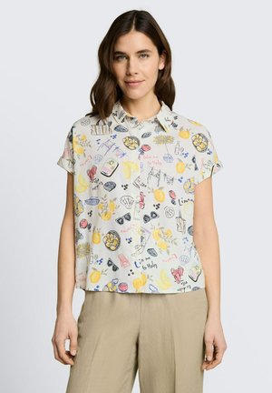 Kobieta w koszuli z krótkim rękawem zapinanej na guziki z kolorowymi, letnimi ilustracjami i beżowych spodniach, stojąca na jednolitym tle.