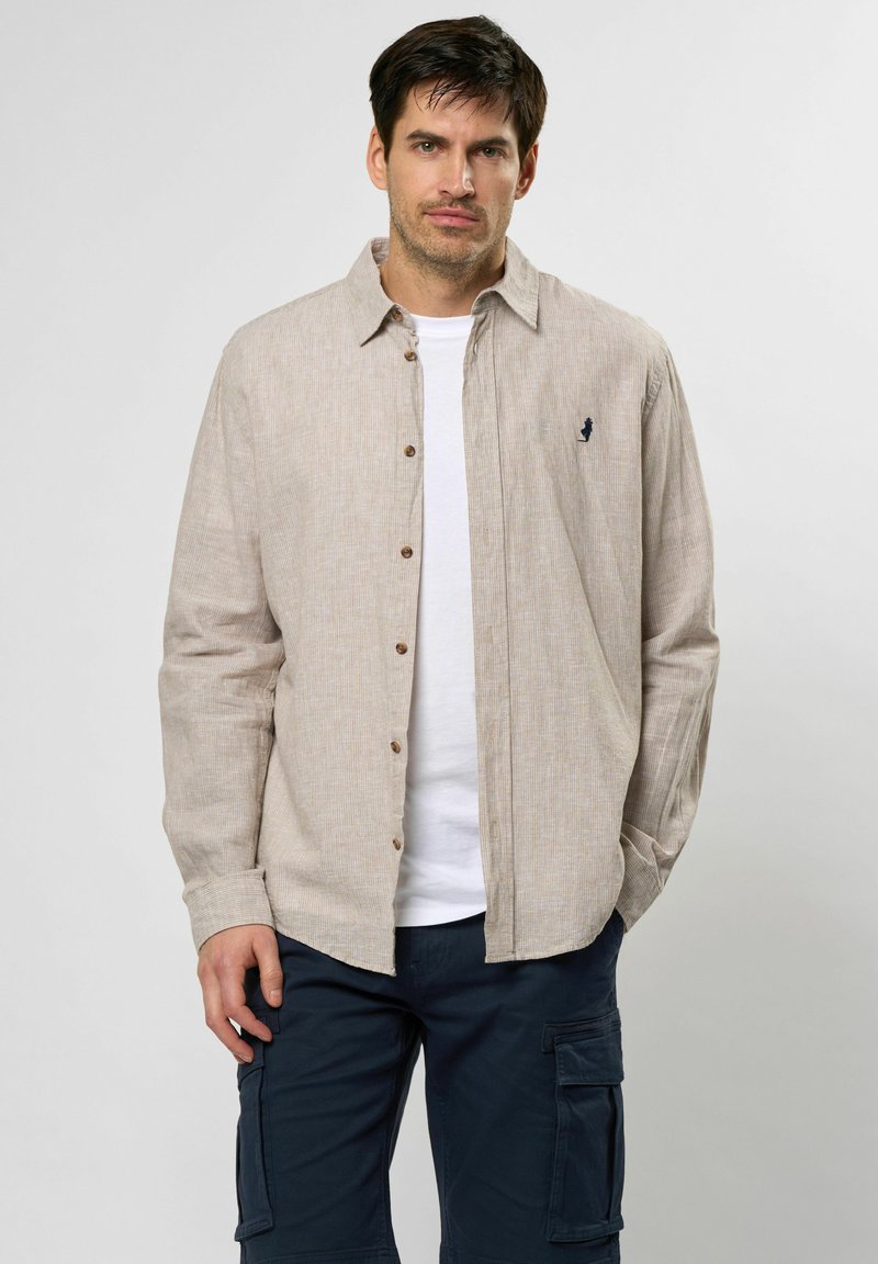 Beige Hemd mit Knopfleiste und strukturiertem Muster sowie einem kleinen Logo auf der Brust; getragen über einem weißen T-Shirt und dunkelblauen Cargo-Hosen.
