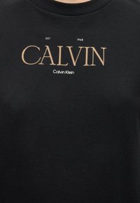 Μαύρο πουκάμισο με μπεζ κείμενο «CALVIN» και λευκό κείμενο «Calvin Klein» από κάτω, και «EST 1968» πάνω από το μεγάλο κείμενο.
