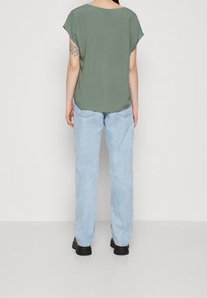 Person iført en olivengrøn kortærmet top, lyseblå jeans, sorte sko, stående med front mod en lys baggrund, og viser en edderkoppespindstatovering på venstre arm.