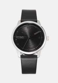 Montre analogique noire avec un cadran noir brossé, des accents argentés, un boîtier en acier inoxydable et un bracelet en cuir noir. Présente des marqueurs d'heure minimalistes.