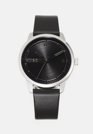 Montre analogique noire avec un cadran noir brossé, des accents argentés, un boîtier en acier inoxydable et un bracelet en cuir noir. Présente des marqueurs d'heure minimalistes.