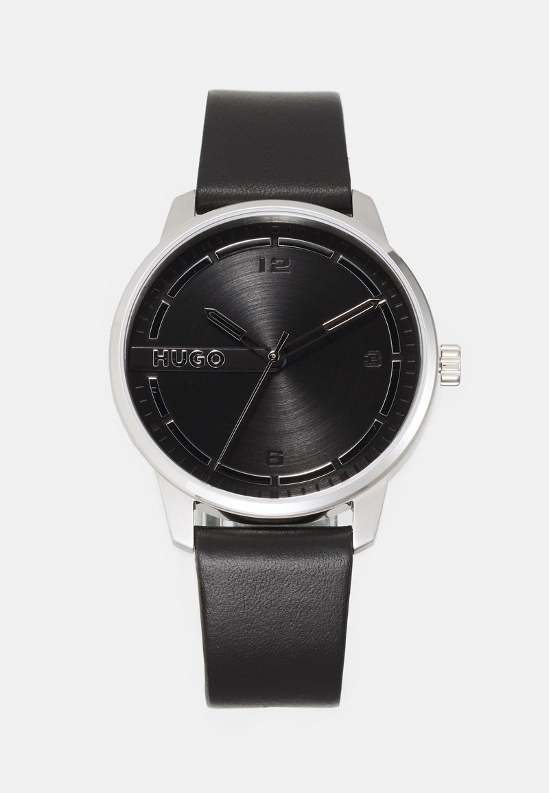 Montre analogique noire avec un cadran noir brossé, des accents argentés, un boîtier en acier inoxydable et un bracelet en cuir noir. Présente des marqueurs d'heure minimalistes.