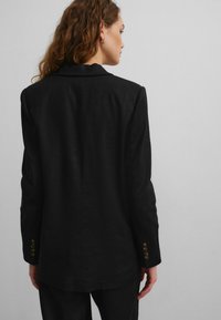 Black blazer made of textured fabric, featuring a single vent at the back and three bronze-button accents on each cuff.