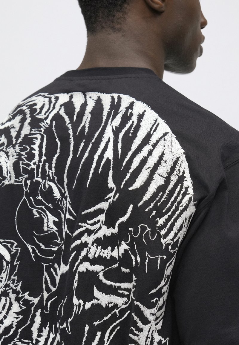 Sweatshirt noir avec un design de tigre brodé en blanc au dos, porté par une personne de dos qui regarde sur le côté.