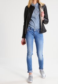 Černá prošívaná bunda přes světle modrou džínovou košili, spárovaná se světle modrými džíny a stříbrnými teniskami. Hladké textury a uvolněný střih.