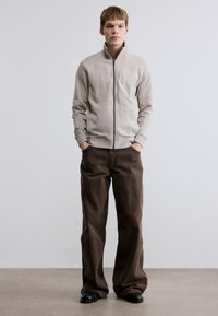 Ανοιχτό γκρι μπουφάν με φερμουάρ από απαλό υλικό, σε συνδυασμό με σκούρο καφέ παντελόνι με φαρδιά μπατζάκια. Υφάσμα με υφή, μίνιμαλ σχεδιασμός, χωρίς ορατές λεπτομέρειες.