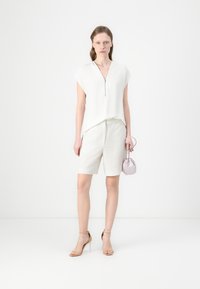 Hvit glidelåsbluse med korte ermer, beige skreddersydde shorts og en lilla rund håndveske. Modellen har på seg nakensandaler med høye hæler.