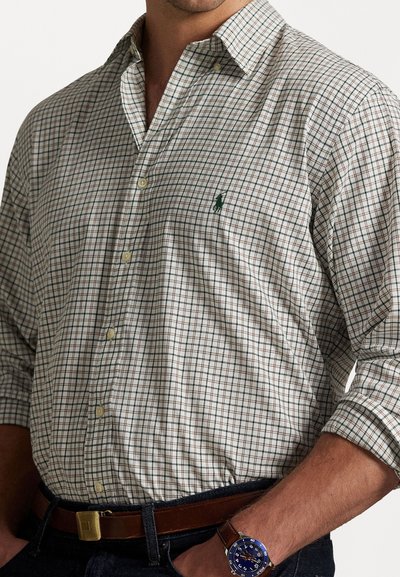 Mies, joka on pukeutunut vaaleanbeigeen ruudulliseen nappipaitaan, jonka hiharat ovat käännettyinä, tummiin farkkuihin, ruskeaan nahkavyöhön ja sinitaustaisiin rannekelloihin, joissa on ruskea hihna.