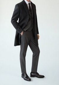 Donker wollen overjas gecombineerd met een grijs geruite vest, wit overhemd en zwarte stropdas. Compleet met gepolijste zwarte leren schoenen.