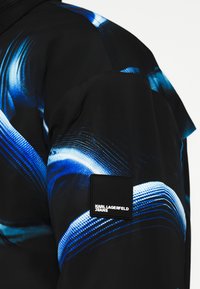 Čierna bunda s modrými abstraktnými vzormi, hladká textura a štvorcová nášivka Karl Lagerfeld Jeans na rukáve.