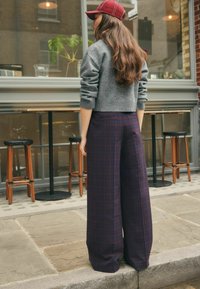 Grijze gebreide trui en brede geruite broek in donkerrood en marineblauw, gecombineerd met een bordeauxrode pet. Buitencafé omgeving met houten krukken.