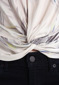 Krátký rukáv top v lehkém materiálu s uzlovým detailem. Obsahuje jemný abstraktní tisk v pastelových barvách. Kombinováno s tmavými džíny a detaily na knoflících.