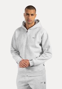 Muž v světle šedé zipové mikině a odpovídajících teplákách, se sepjatýma rukama před sebou, stojící na jednoduchém světlém pozadí.