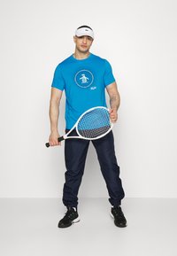 Blå t-skjorte med korte ermer og sirkulært logo-print, sammen med marineblå joggebukser og svarte treningssko, mens han holder et hvitt tennisracket.