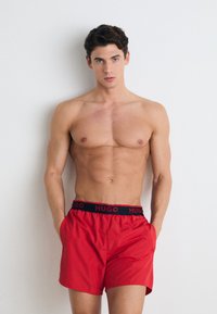 Shorts de bain rouges avec une taille noire arborant le texte "HUGO". Le matériau semble lisse et léger, avec une coupe ample et des poches latérales.