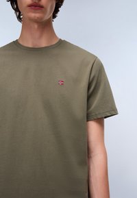 Oliven grønn bomulls T-skjorte med korte ermer, med en liten brodert flaggdetalj på brystet og rund hals.