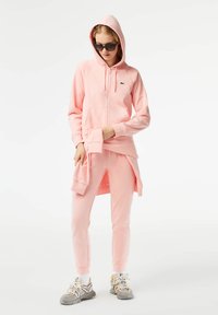 Roza zip-up dukserica s kengurudžem, u kombinaciji s kapri hlačama. Lagani materijal, opušteni kroj, rebrasti manžeti i logo detalj.