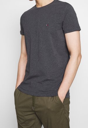 T-shirt gris foncé à manches courtes en tissu doux, arborant un petit logo rouge, blanc et bleu sur la poitrine. Associé à un pantalon vert olive.