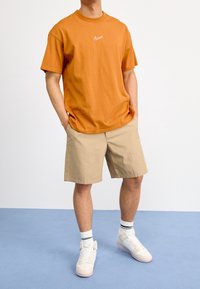 Henkilö, jolla on päällä oranssi Nike-t-paita, vaaleat shortsit, valkoiset Nike-tennarit ja valkoiset sukat, joissa on mustat raidat, seisoo sinisellä lattialla.