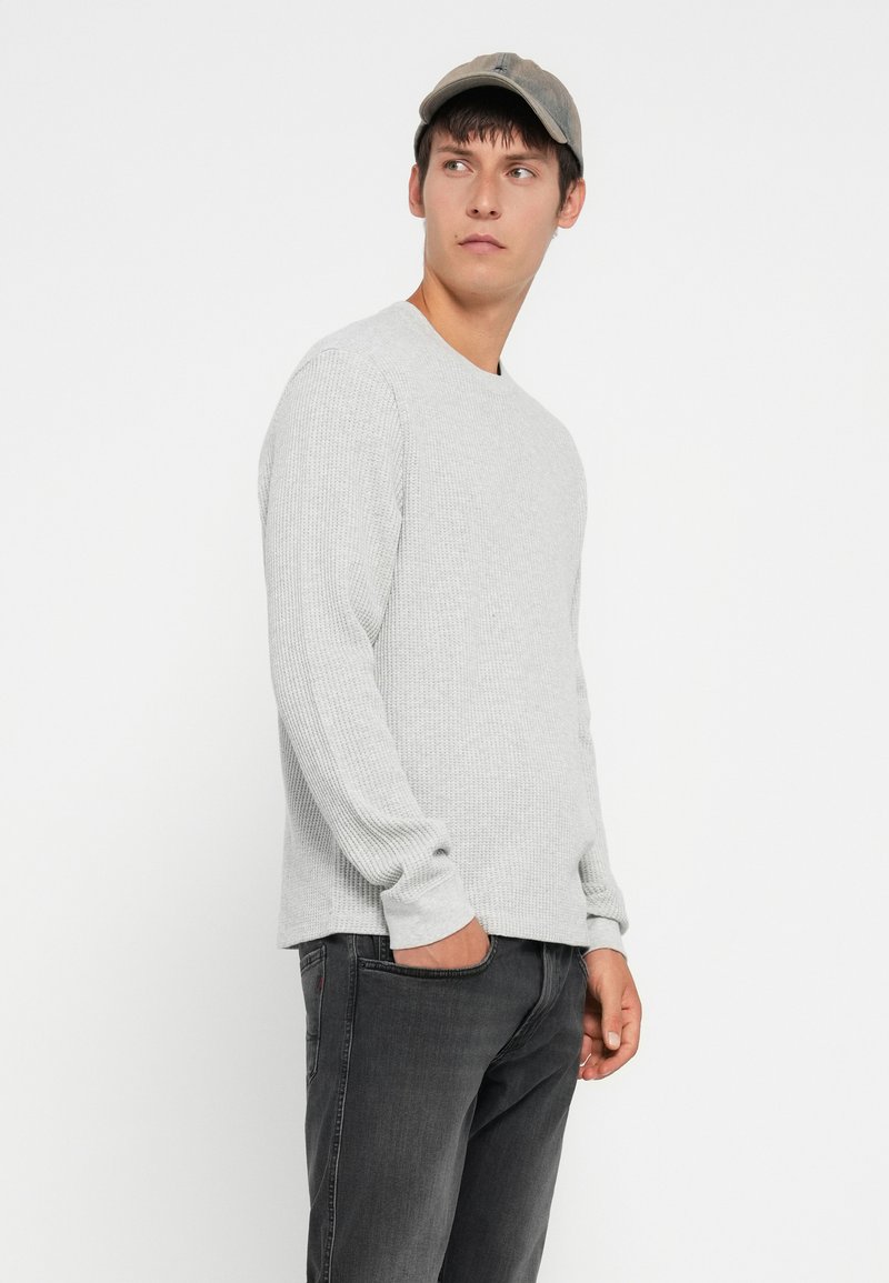 Lichtgrijs langmouw geribbelde trui met een ronde hals, gecombineerd met donkergrijze jeans en een beige pet.