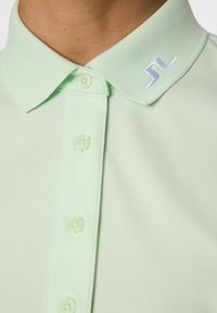 Vihreä kauluspaita, jossa on tekstuuroitu kaulus, kuusi nappia ja valkoisella brodeerattu logo kauluksessa. Sileä kangasviimeistely.