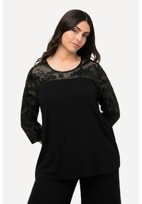Schwarzes Oberteil mit transparenter Spitze und strukturiertem Muster sowie Dreiviertelärmeln, kombiniert mit schwarzen Hosen.