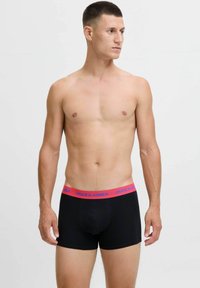 Mlad moški s kratkimi lasmi, oblečen v črne boksarice z rdečim pasom, na katerem piše "JACK & JONES," stoji ob preprosti beli ozadju.