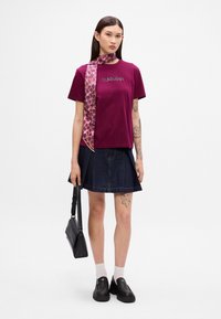 Calvin Klein burgunder t-skjorte med fet logo, sammen med et denimskjørt. Tilbehør med et mønstrete skjerf og en svart crossover-veske.