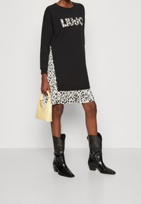 Schwarzes Sweatshirt-Kleid mit "Liu Jo"-Druck, ausgestattet mit einem weißen, tierisch gemusterten Volantabschluss und langen Ärmeln. Begleitet von einer gelben Handtasche.