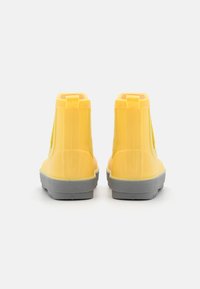 Dzelteni gumijas potīšu zābaki ar gludu virsmu, strukturētām pelēkām zolēm un vilkšanas cilpiņām aizmugurē vieglai novilkšanai.