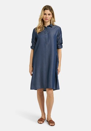 Frau trägt ein knielanges blaues Jeans-Hemdkleid mit hochgekrempelten Ärmeln und braunen flachen Sandalen und steht auf einem schlichten hellen Hintergrund.