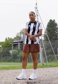 Schwarz-weißer gestreifter Fußballtrikot mit Adidas-Logo, kombiniert mit einem braunen Rock. Weiße Sportschuhe und Socken bis zum Knöchel. Hintergrund mit Gras und Torpfosten.