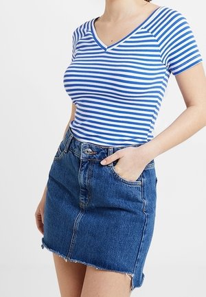 Kobieta ubrana w niebiesko-biały paskowany top z krótkim rękawem i dekoltem w serek, włożony do wysokiej taliowanej spódnicy jeansowej z surowym wykończeniem, z ręką w kieszeni.