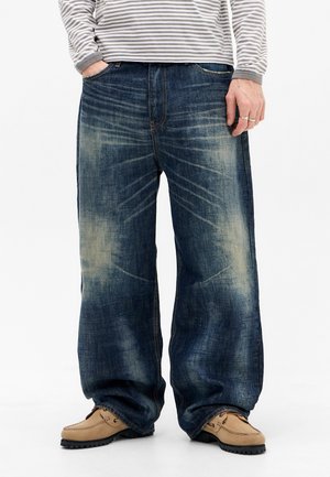 Person trägt weit geschnittene ausgebleichte blaue Jeans, beige Mokassinschuhe und ein grau-weiß gestreiftes langärmliges Shirt, Hände teilweise sichtbar.
