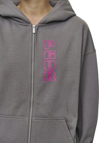Felpa grigia con zip e cappuccio, caratterizzata da grandi lettere rosa "PGDR" sul davanti. Realizzata in un morbido tessuto testurizzato con due tasche frontali.