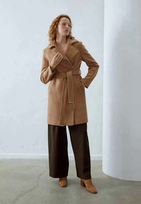 Manteau couleur camel avec ceinture, col cranté, détails de coutures verticales et coupe ajustée, associé à un pantalon large marron et des talons.