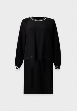 Une femme porte une robe noire longueur genou avec des bordures rayées et des bottines noires, sur un fond clair uni.