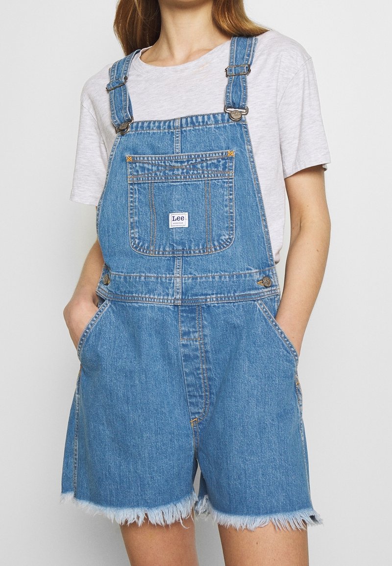 Personne portant une salopette courte en jean bleu avec une poche avant étiquetée "Lee" sur un t-shirt à manches courtes gris clair, les mains dans les poches.