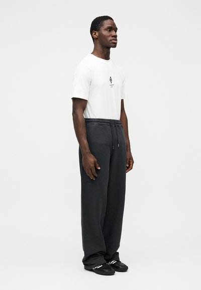 Fehér póló kis fekete grafikával az elején, laza fekete melegítőnadrággal párosítva. A modell oldalt áll, fekete cipőt visel.