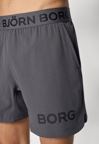 Szürke atletikus rövidnadrág rugalmas derékpánttal, amelyen a „BJÖRN BORG” felirat olvasható, valamint a „BORG” nyomtatás a szegély közelében, egy személy combközép magasságában.