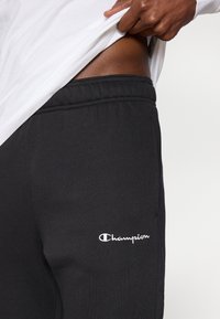 Μαύροι παντελόνια jogger από φλις με ελαστική μέση και λευκό κεντημένο λογότυπο "Champion" σ το πλάι.